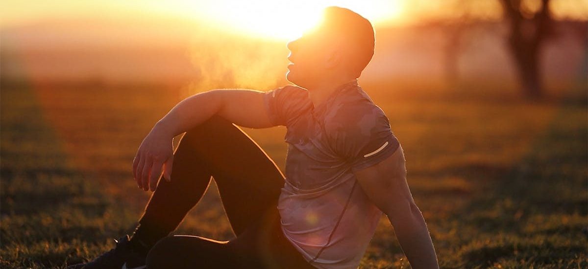 Muscular man sits in rural field at sunset, exhaling visible breath.