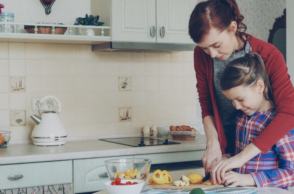 Young pretty mother teaching her cute daughter to cut vegetables properly. Little smiling girl cooking together with loving mom at home in modern kithcen