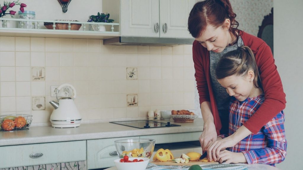 Young pretty mother teaching her cute daughter to cut vegetables properly. Little smiling girl cooking together with loving mom at home in modern kithcen