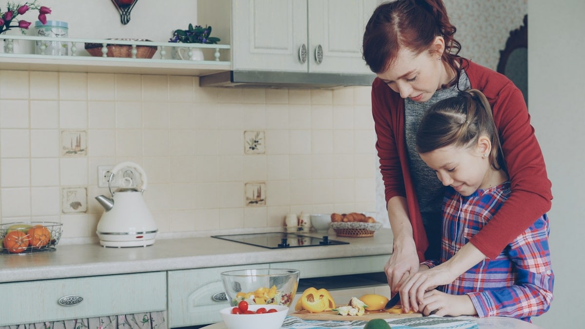 Young pretty mother teaching her cute daughter to cut vegetables properly. Little smiling girl cooking together with loving mom at home in modern kithcen
