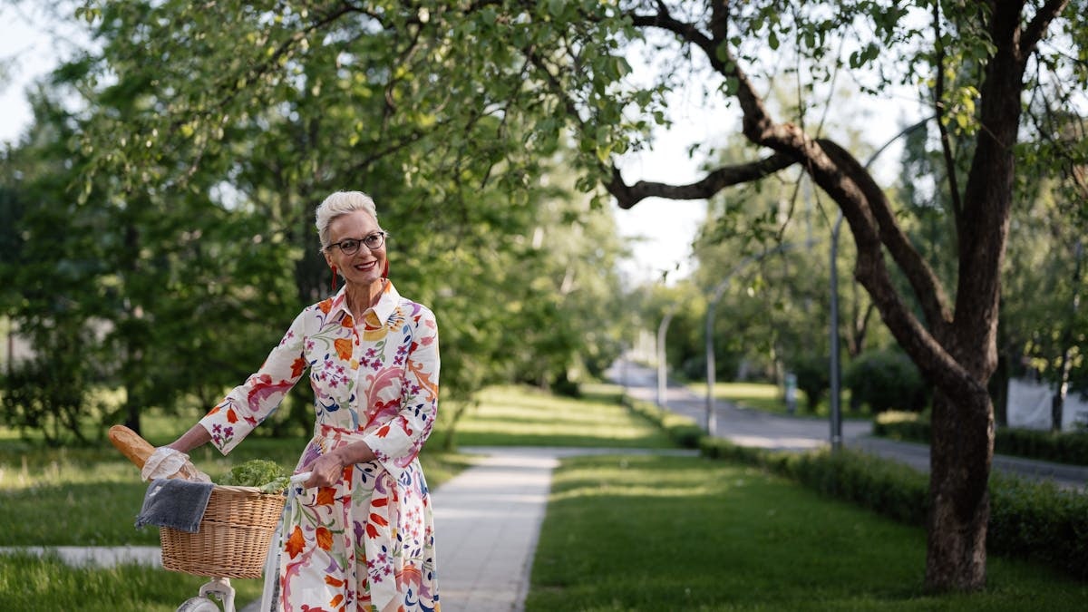 Smiling senior woman in floral dress walking bicycle outdoors on a sunny day.